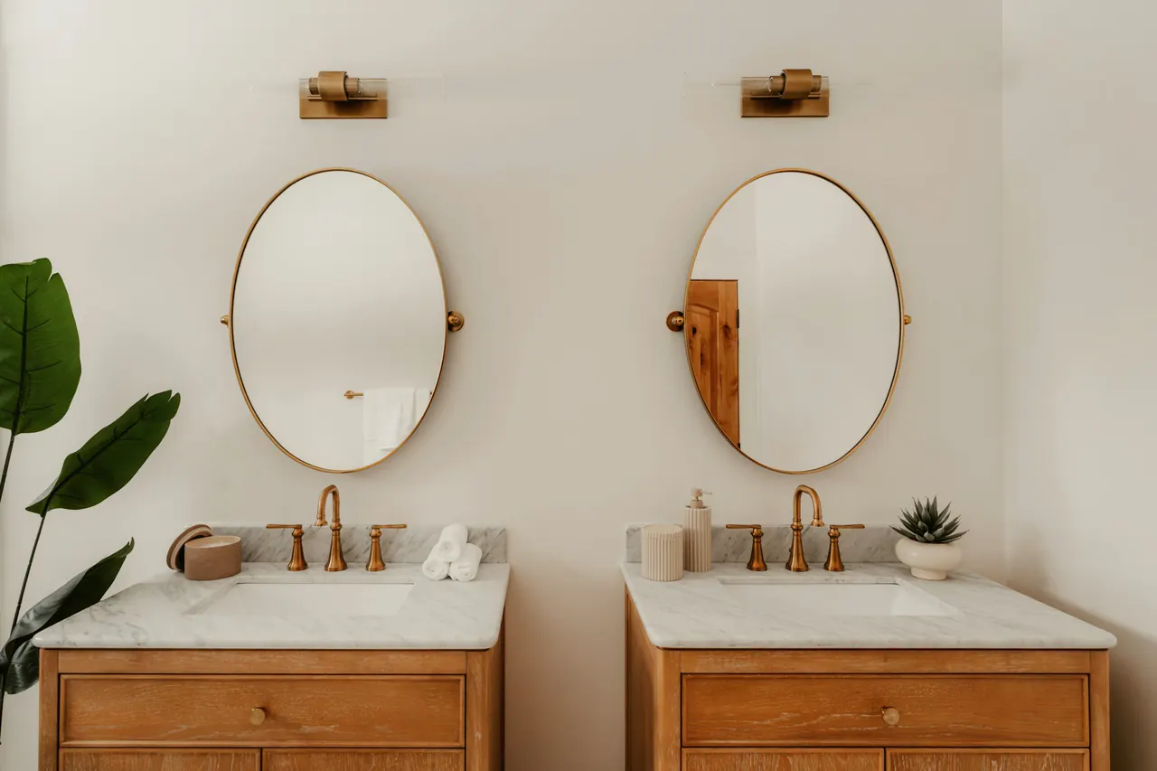 Two wooden bathroom vanities with oval mirrors and gold fixtures